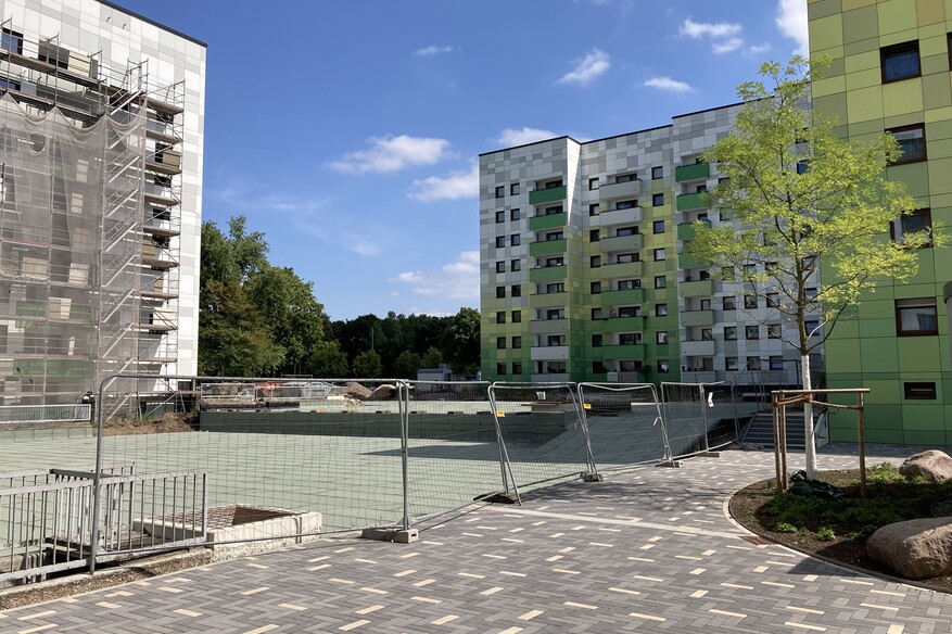 Blick auf grüne Gebäude, davor eine gepflasterte Fläche, die von einem Bauzaun getrennt wird.