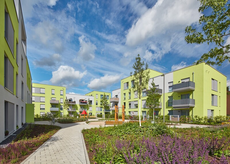 Blick in der Quartier, links und im Hintergrund grün-weiße Gebäude. Vorne Blumenbeete und Wege.