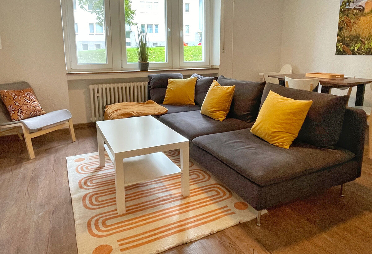 Ein Wohnzimmer mit L-förmigem, Sofa, Couchtisch und Teppich. Ein Sessel steht links, rechts hinten ein Esstisch.