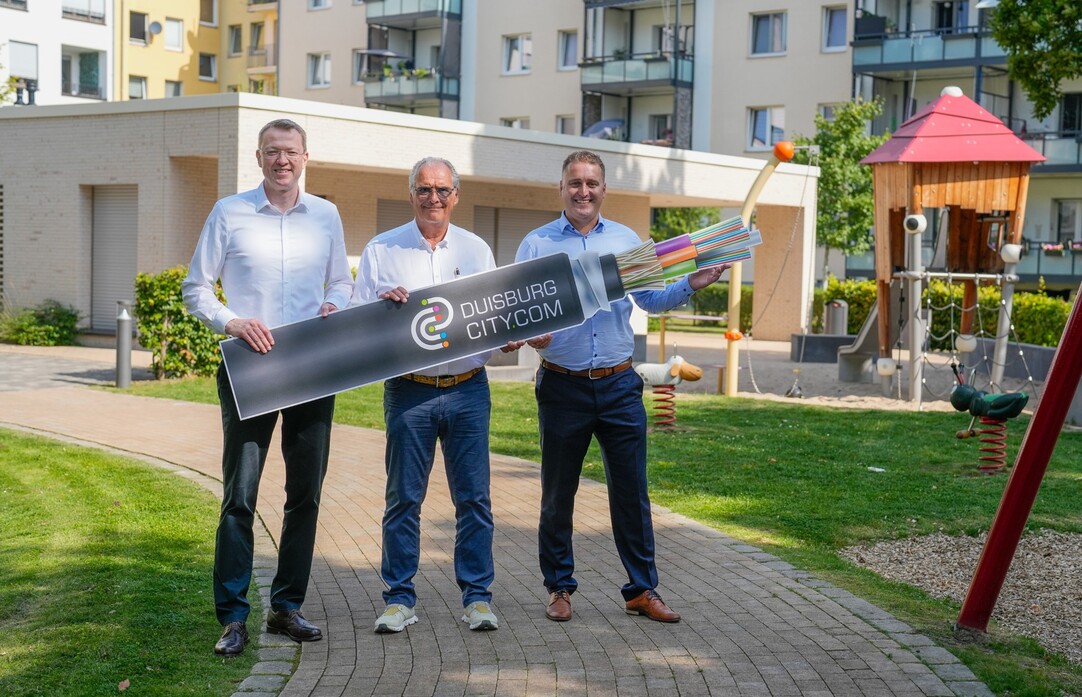 Drei Männer stehen in einem Innenhof mit Spielplatz und Wiesen und halten ein Pappschild hoch, das ein Glasfaserkabel zeigt.