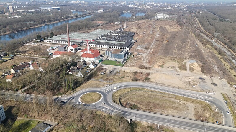 Luftbildaufnahme einer Fläche mit einem Kreisverkehr und Straßen im Vordergrund. Dahinter alte Fabrikgebäude mit grauen Dächern, eines mit rotem Schornstein.