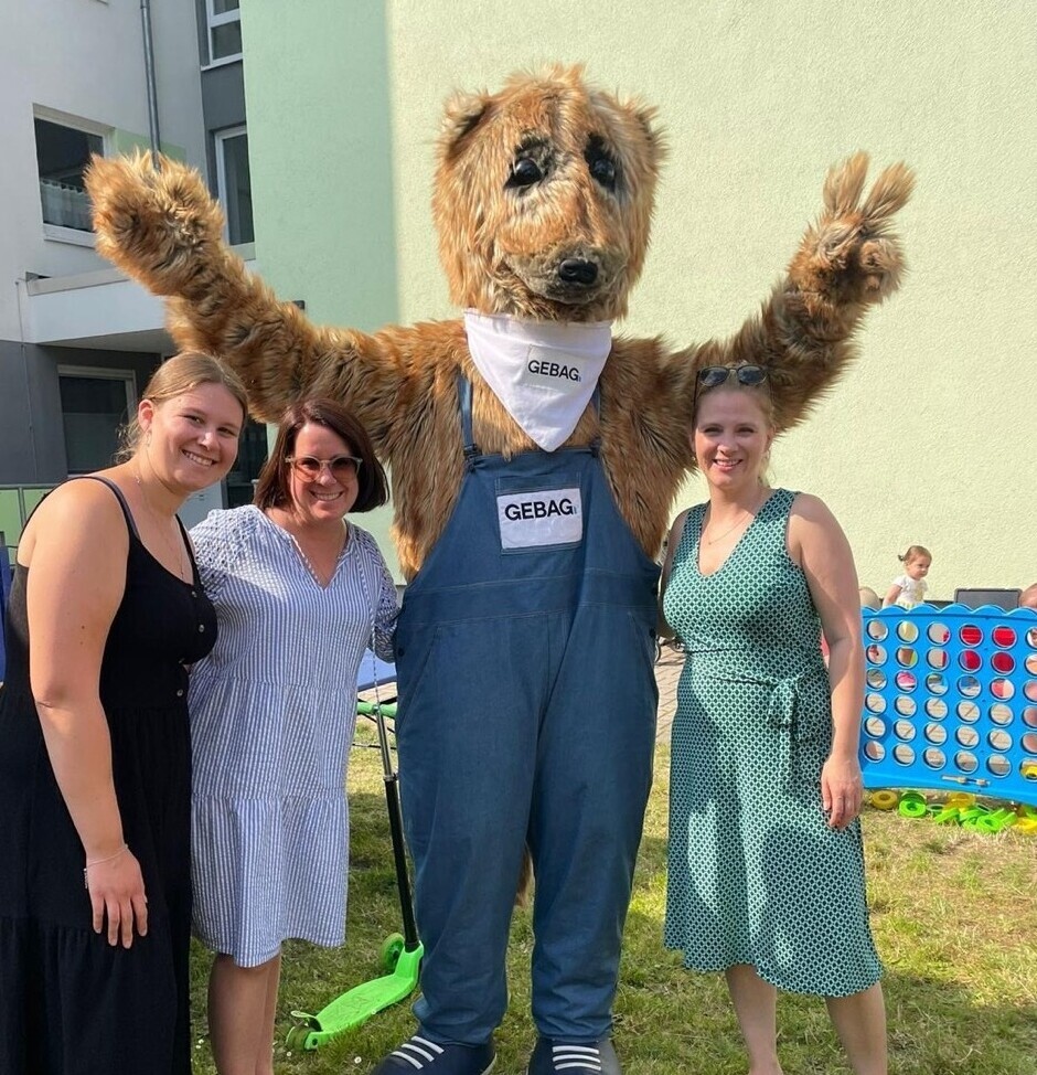 Drei Frauen posieren mit einem Erdmännchen-Maskottchen vor einem Wohnhaus.