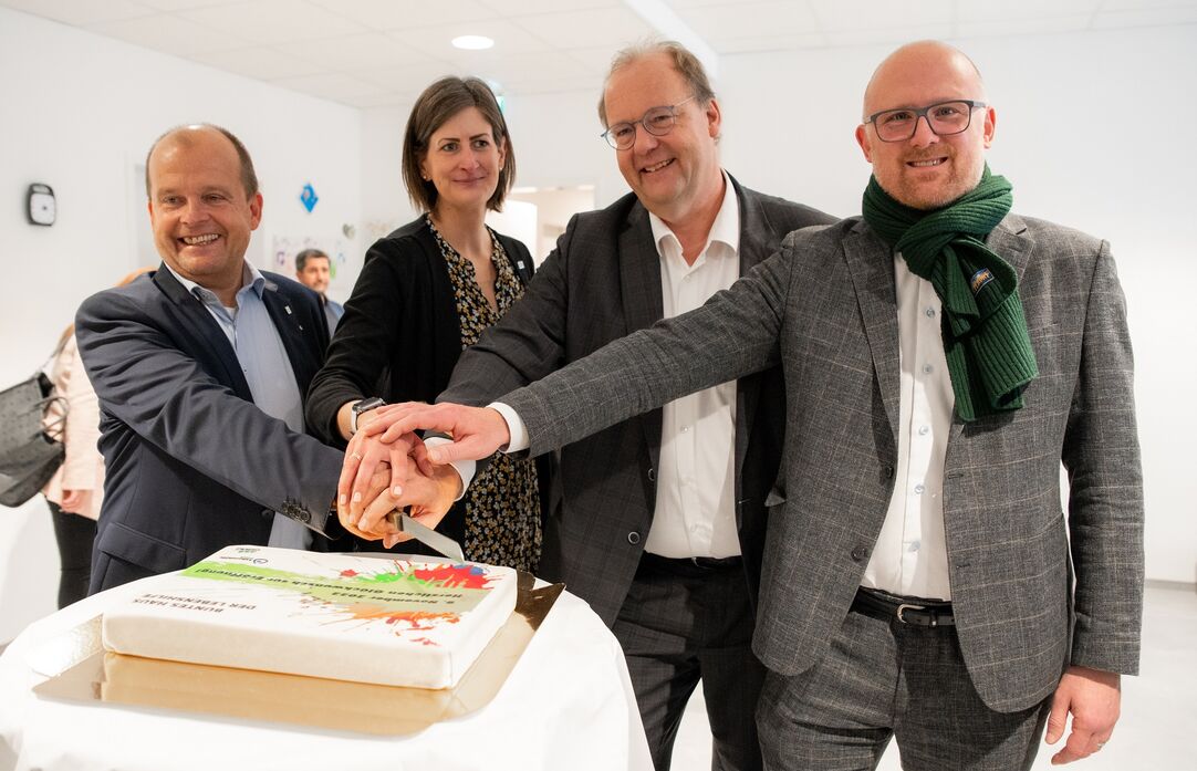 Vier Menschen (eine Frau, drei Männer) schneiden gemeinsam eine Torte an und lächeln dabei.