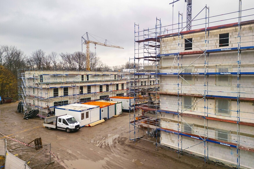 Blick auf zwei eingerüstete Mehrfamilienhäuser auf einer Baustelle. Zwischen den Häusern Container und Fahrzeuge. Der Boden ist sandig.