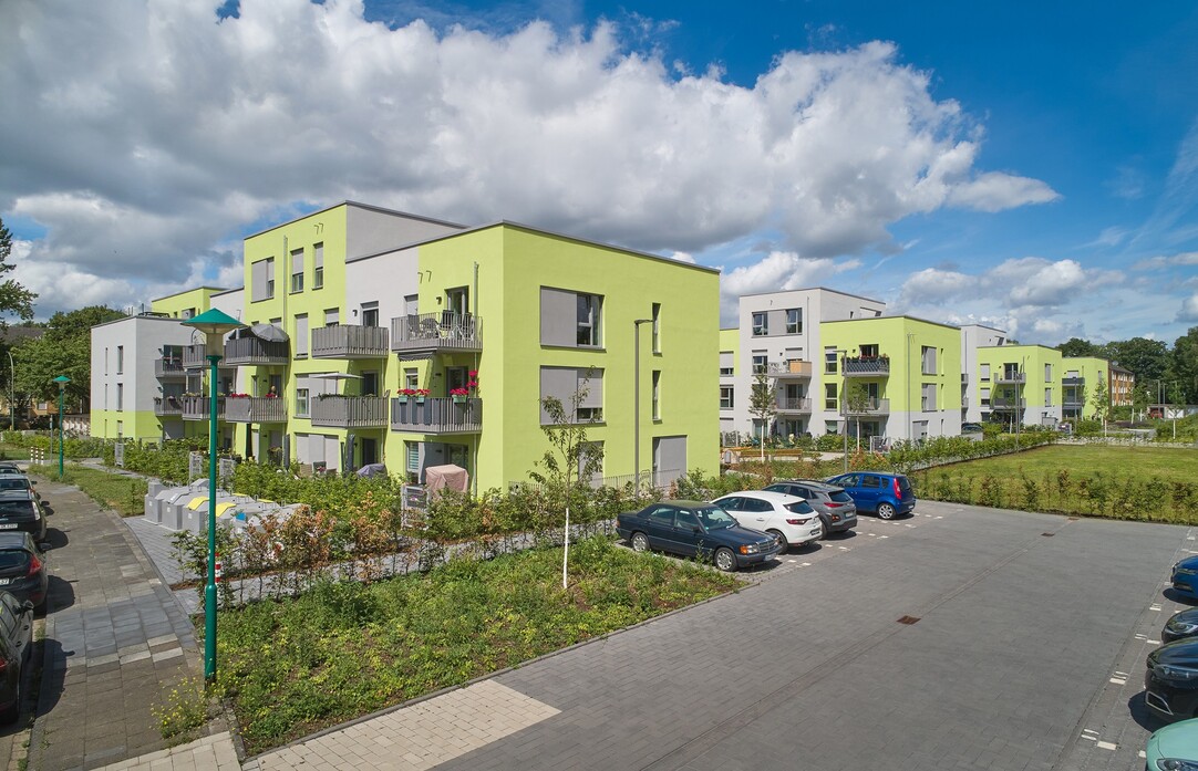 Ansicht des neuen Quartiers mit grün-weißen Gebäuden. Vorne rechts ein Parkplatz mit einigen Autos.