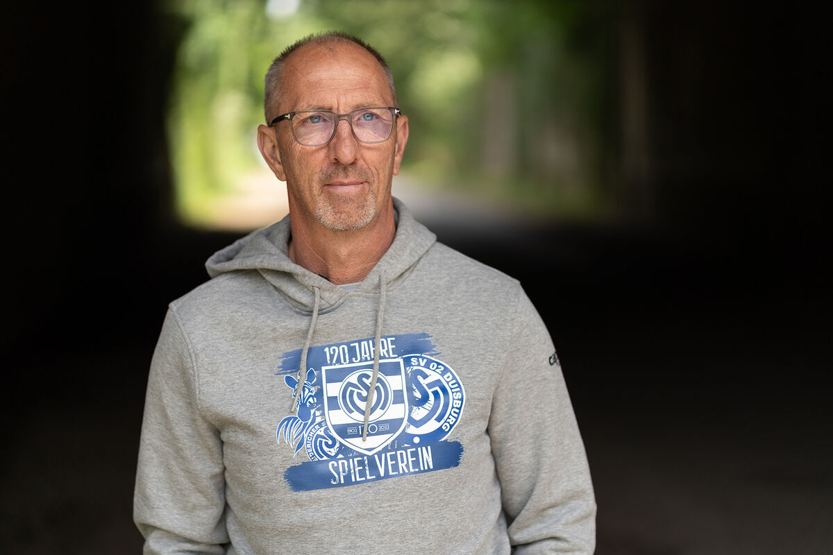 Ein Mann mit kurzen grauen Haaren, Bart und Brille trägt einen grauen Kapuzenpulli mit einem blauen MSV Logo darauf.