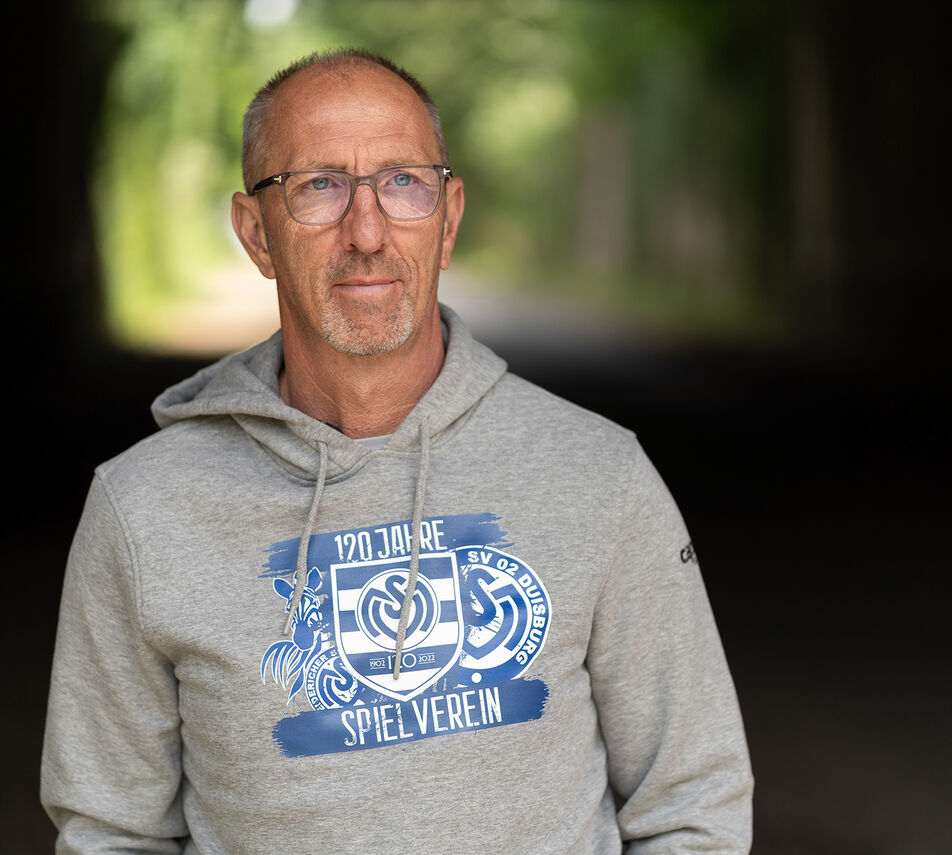 Ein Mann mit kurzen grauen Haaren, Bart und Brille trägt einen grauen Kapuzenpulli mit einem blauen MSV Logo darauf.