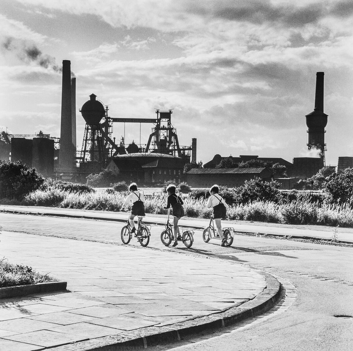 Eine historische Schwarz-Weiß-Aufnahme zeigt 3 Kinder auf Fahrrädern. Dahinter steht eine große Industrieanlage.