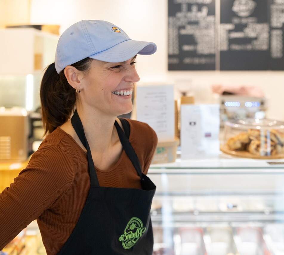 Eine lächelnde junge Frau mit hellblauem Basecap und schwarzer Schürze steht vor einer Theke in einem Eiscafé.