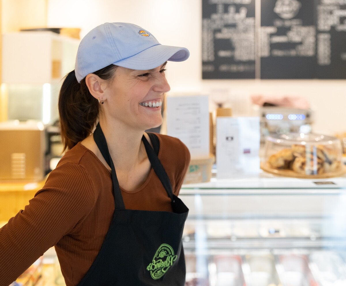 Eine lächelnde junge Frau mit hellblauem Basecap und schwarzer Schürze steht vor einer Theke in einem Eiscafé.