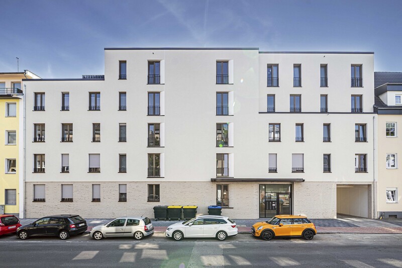Straßenansicht des Neubaus in der Grabenstraße, davor sind Autos geparkt. Die Fassade ist unten in sandfarbenen Steinen gefasst, darüber hell verputzt.