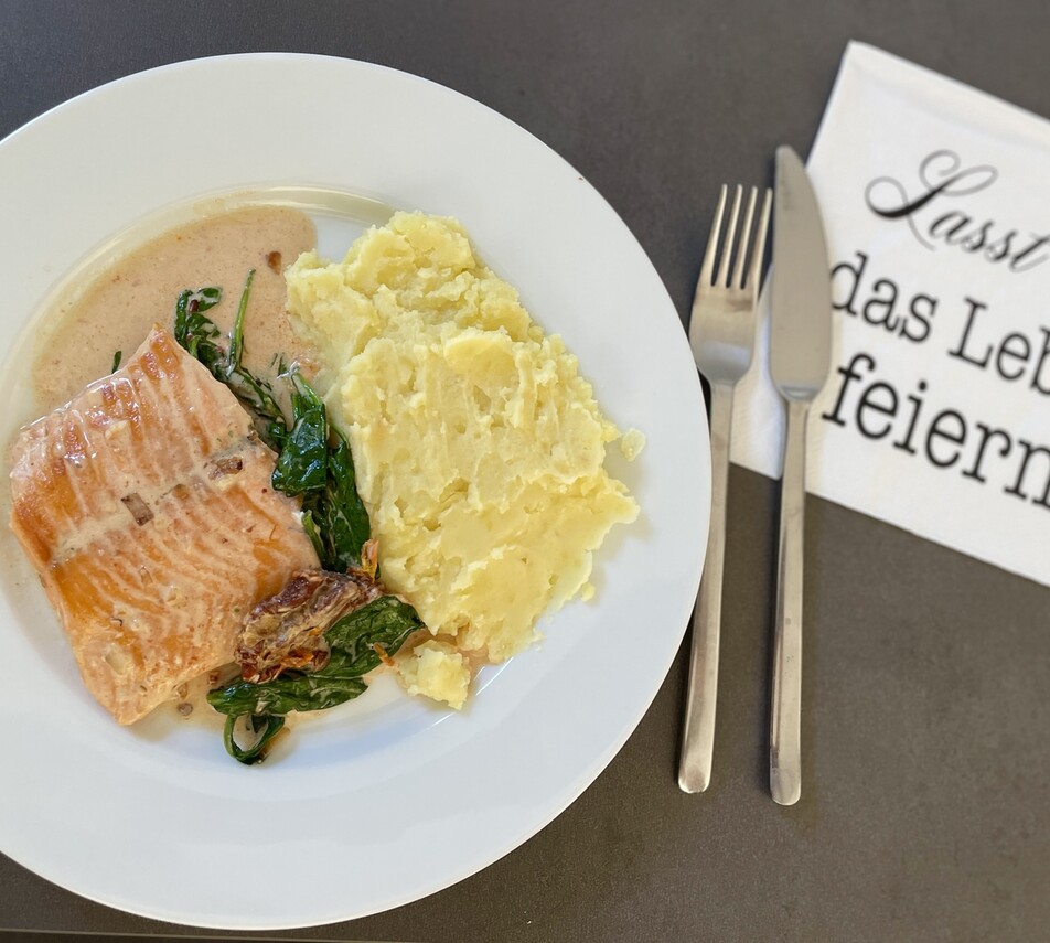 Ein Teller mit Lachsfilet, Kartoffelpürree, Sauce und Spinat, daneben Besteck und Serviette