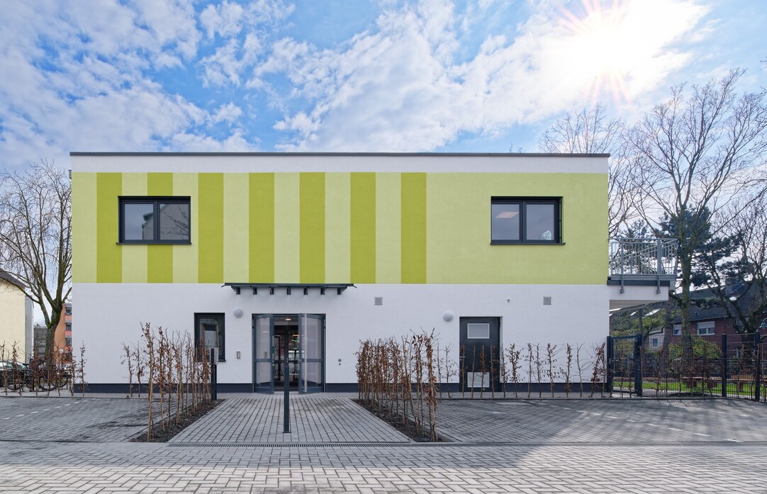 Außenansicht der Kita, Fassade oben hellgrün, unten weiß. Vor dem Gebäude Parkplätze und junge Hecken. Himmel hellblau mit Wolken