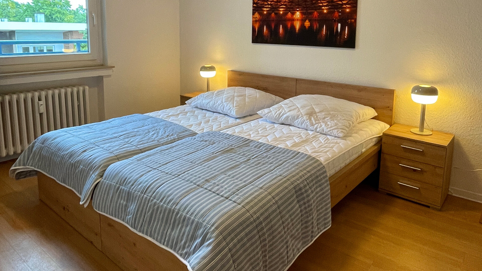 Ein Doppelbett mit Nachttischen und Lampen steht in einem Schlafzimmer mit Holzboden und Fenster.