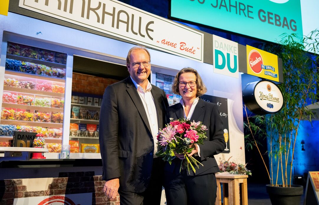 Ein Mann und eine Frau mit einem Blumenstrauß stehen vor einer Trinkhallenkulisse.