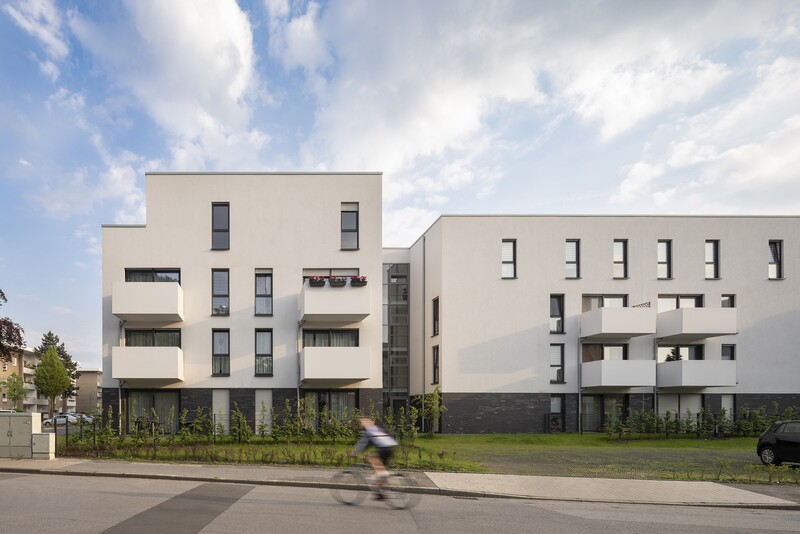 Ansicht des Mehrfamilienhauses mit Balkonen und Fenstern, davor Rasen und Straße. Schemenhaft ist ein Radfahrer zu erkennen.