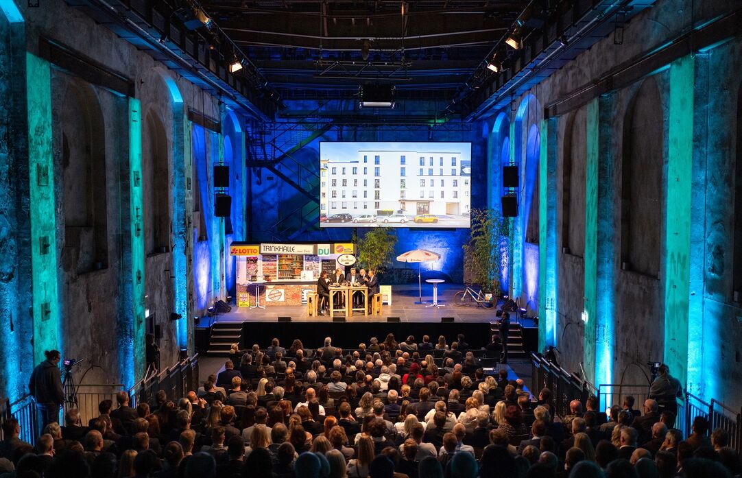 Eine große Menschenmenge sitzt in einer Halle und schaut auf eine Bühne mit Rednern und einer Leinwand.