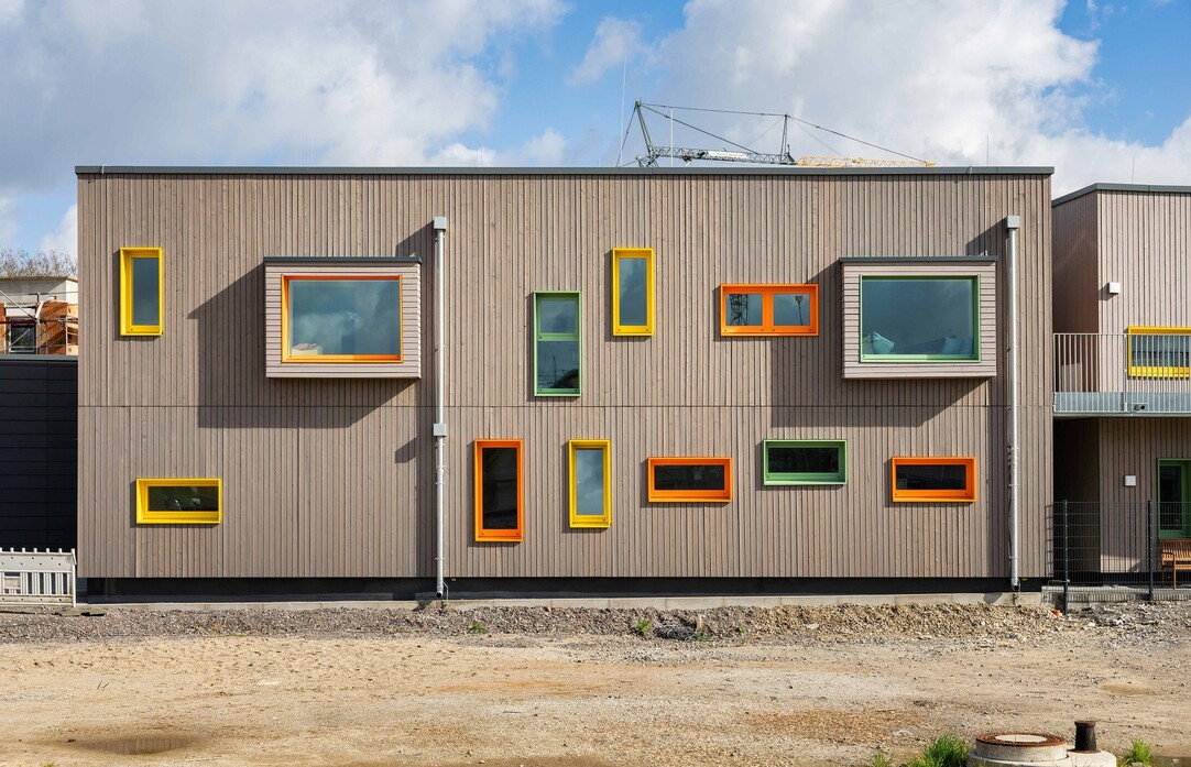 Holzfassade der Kita mit vielen Fenstern in unterschiedlichen Größen und Formaten, alle mit farbigen Rahmen.