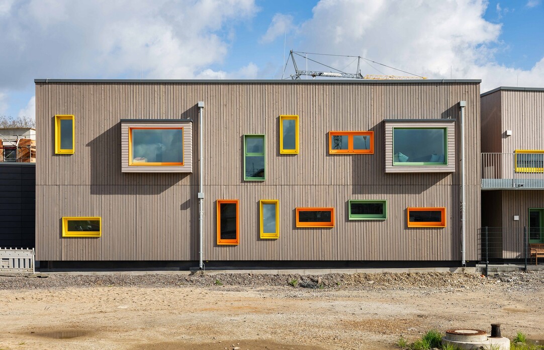 Holzfassade der Kita mit vielen Fenstern in unterschiedlichen Größen und Formaten, alle mit farbigen Rahmen.
