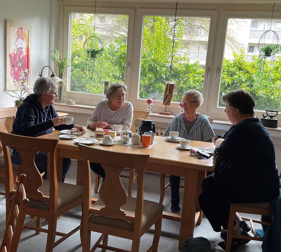 Vier ältere Damen sitzen an einem Holztisch in einem hellen Raum und unterhalten sich angeregt bei Kaffee und Kuchen.