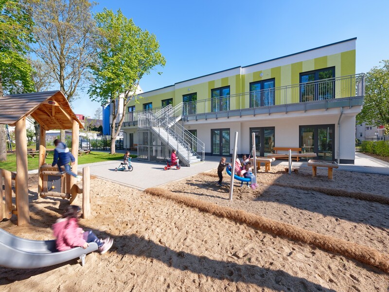 Spielplatz der Kita mit Sandfläche und Geräten, im Hintergrund das grün-weiße Gebäude mit Flachdach.