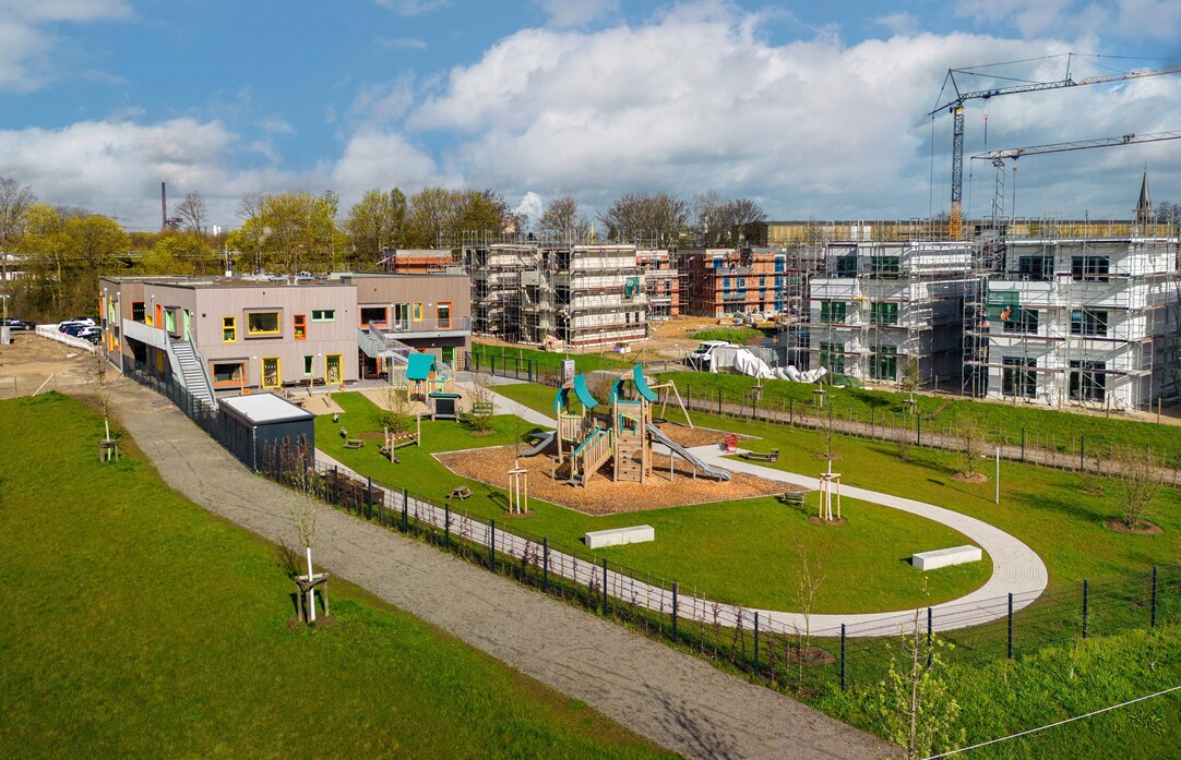 Aufnahme vom schräg oben - Spielplatz mit Rasen und Kita, im Hintergrund das Neubaugebiet Am Alten Angerbach, mit Häusern im Bau.