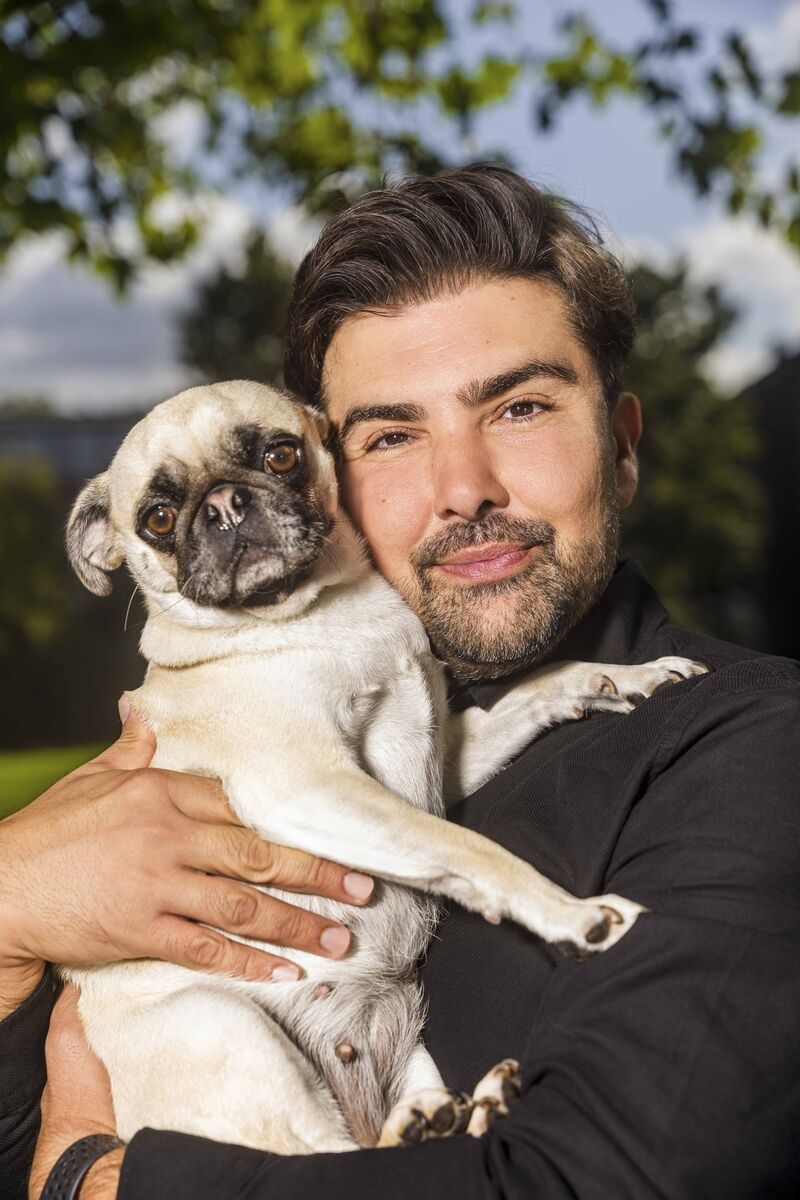 Ein Mann mit kurzen schwarzen Haaren und Bart hält liebevoll einen beigefarbenen Hund in seinen Armen.