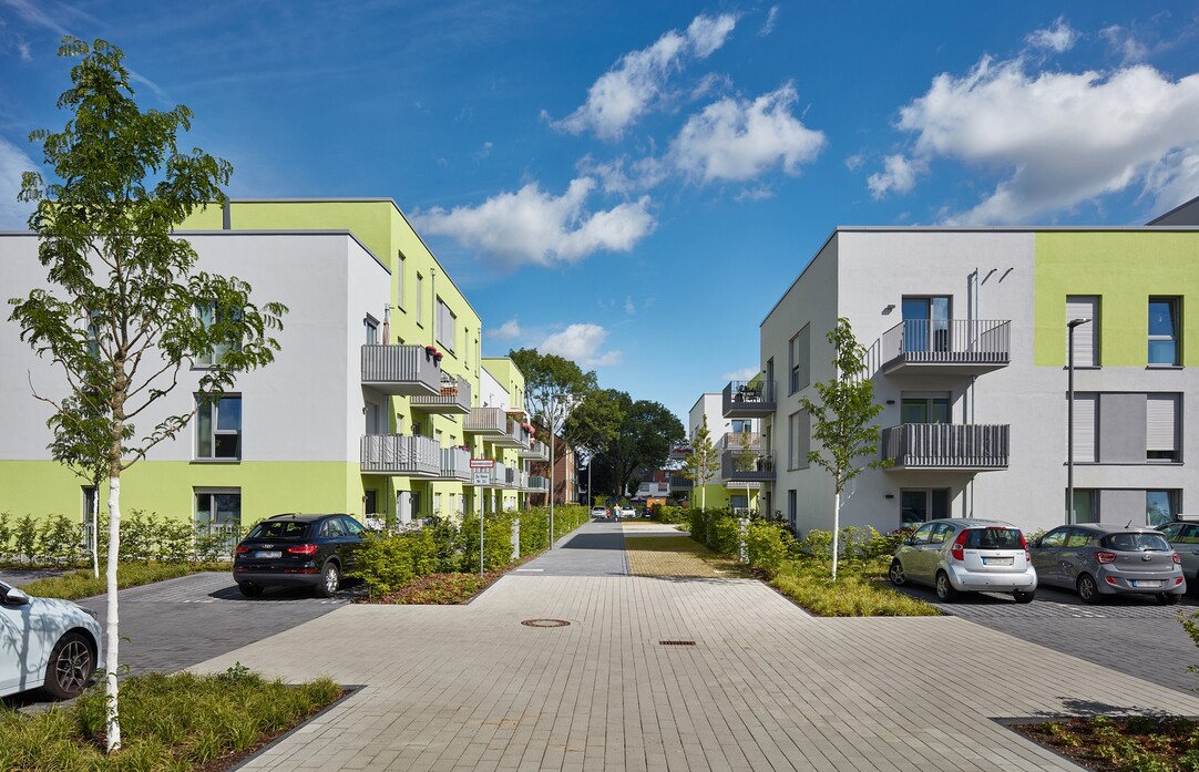 Blick in einen breiten Durchgang im Quartier, links und rechts Häuser. Vorne Parkflächen mit Bäumen.