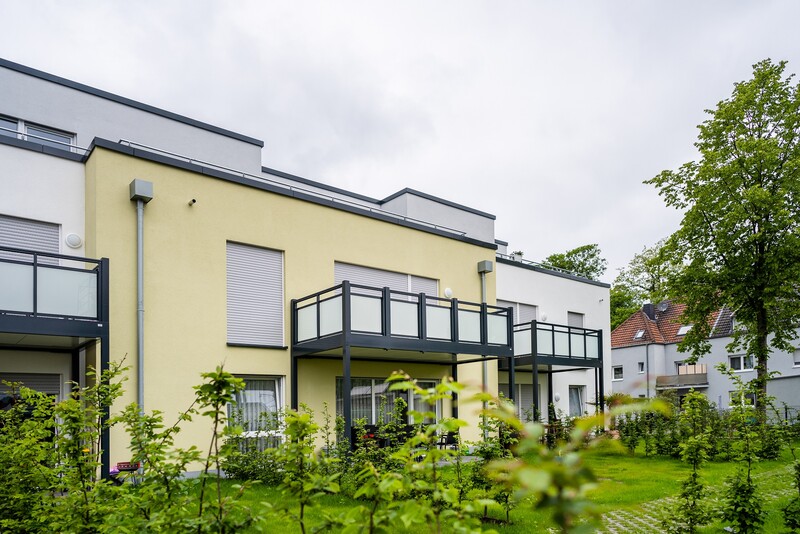 Blick auf ein gelbes Haus mit einem schwarzen Metallbalkon und Milchglasbrüstung. Rechts im Bild ein Baum, vorne eine Rasenfläche.