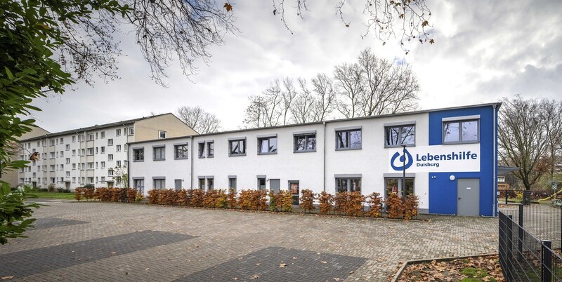 Fronansicht des Gebäudes, ein Teil der Fassade dunkelblau mit Lebenshilfe-Logo. Vor dem Gebäude Parkplätze.