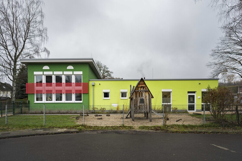 Blick von der Straße auf die Kita, davor ein Zaun und Spielgeräte. Linker Gebäudeteil ist höher, grüne Fassade; rechts niedriger Gebäudeteil mit gelber Fassade.