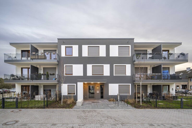 Frontaler Blick auf den Neubau, mittig der Eingangsbereich. Rechts und links kragen Balkone aus. Fassade weiß mit grauen Akzenten (quer).