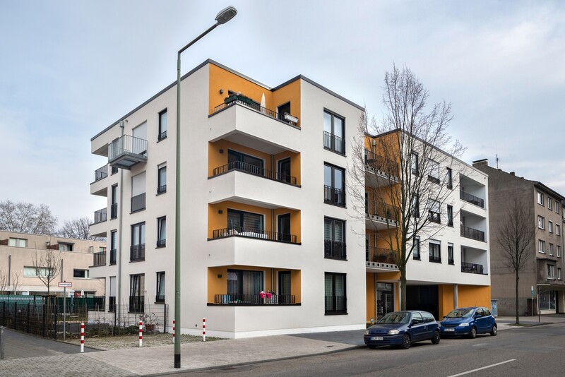 Blick auf die Ecke eines dreistöckigen Hauses. Fassade weiß, mit orangenen Flächen/Akzenten. Zurückgesetzte Balkone. An der Ecke des Hauses eine Laterne.