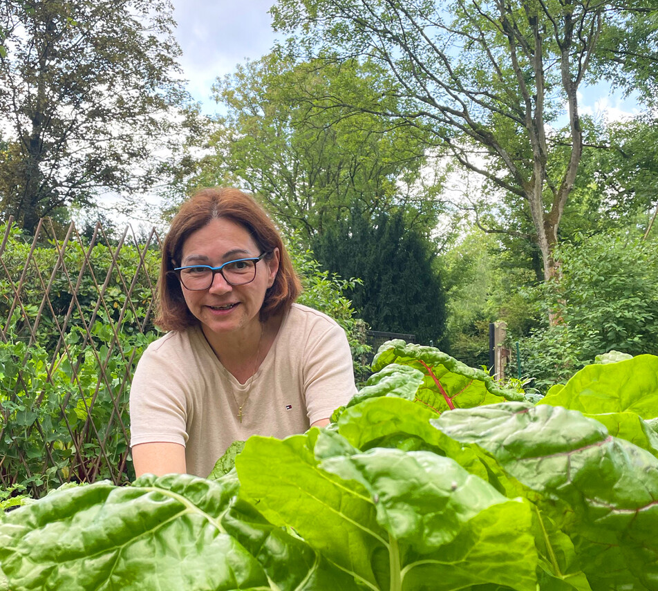 Eine lächelnde Frau mit Brille und schulterlangem, braunem Haar gärtnert in einem Hochbeet.