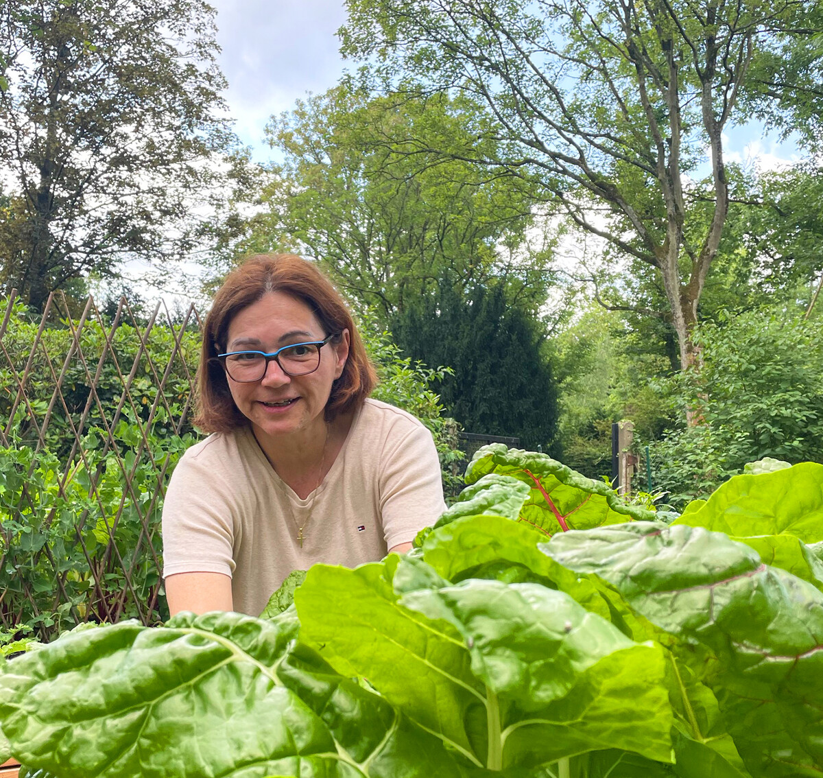 Eine lächelnde Frau mit Brille und schulterlangem, braunem Haar gärtnert in einem Hochbeet.