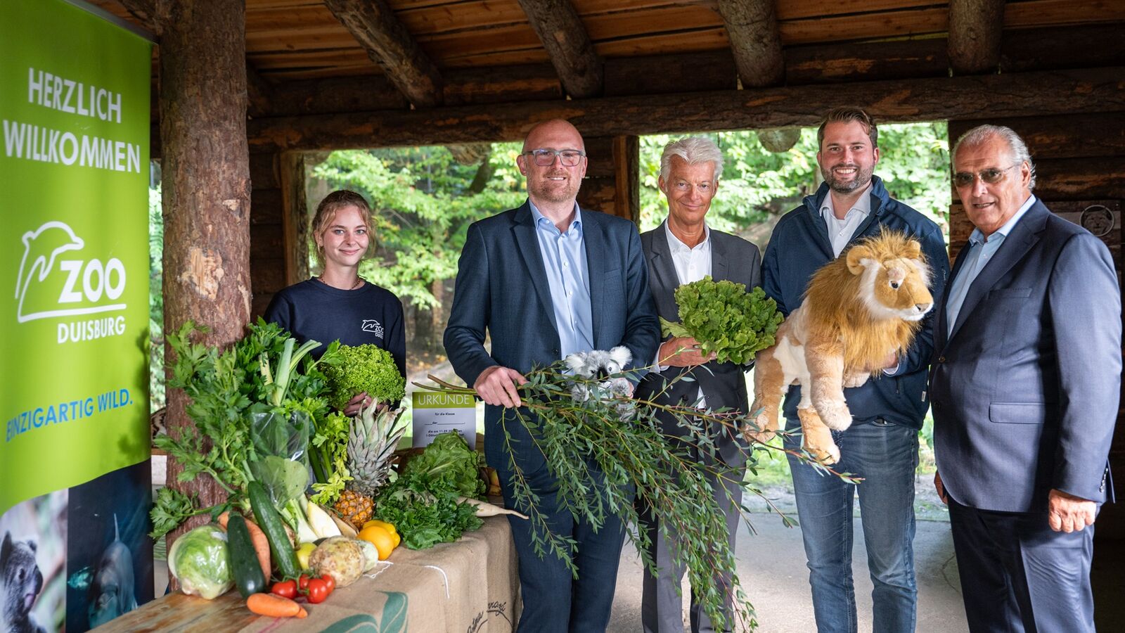 Eine Frau und vier Männer stehen nebeneinander. Sie halten Gemüse und Kuscheltiere in Ihren Händen. Links von Ihnen befindet sich ein Tisch auf welchem ebenfalls Gemüse liegt. Hinter dem Tisch befindet sich ein Banner des Duisburger Zoos.