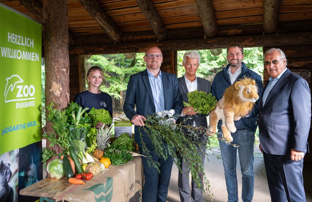 Eine Frau und vier Männer stehen nebeneinander. Sie halten Gemüse und Kuscheltiere in Ihren Händen. Links von Ihnen befindet sich ein Tisch auf welchem ebenfalls Gemüse liegt. Hinter dem Tisch befindet sich ein Banner des Duisburger Zoos.