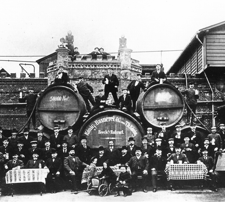 Eine alte Schwarz-Weiß-Fotografie zeigt viele Männer sowie einige Frauen und Kindern vor Bierfässern einer Brauerei