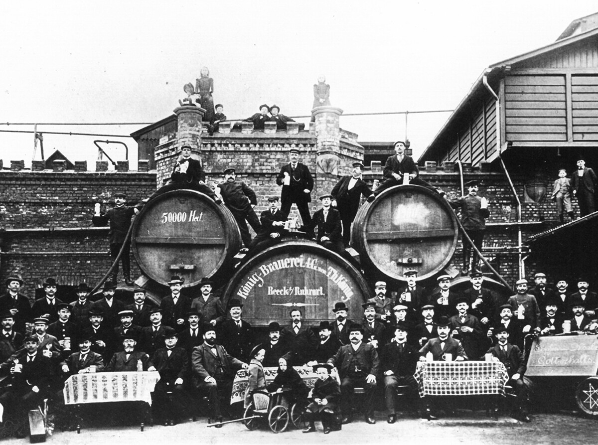 Eine alte Schwarz-Weiß-Fotografie zeigt viele Männer sowie einige Frauen und Kindern vor Bierfässern einer Brauerei