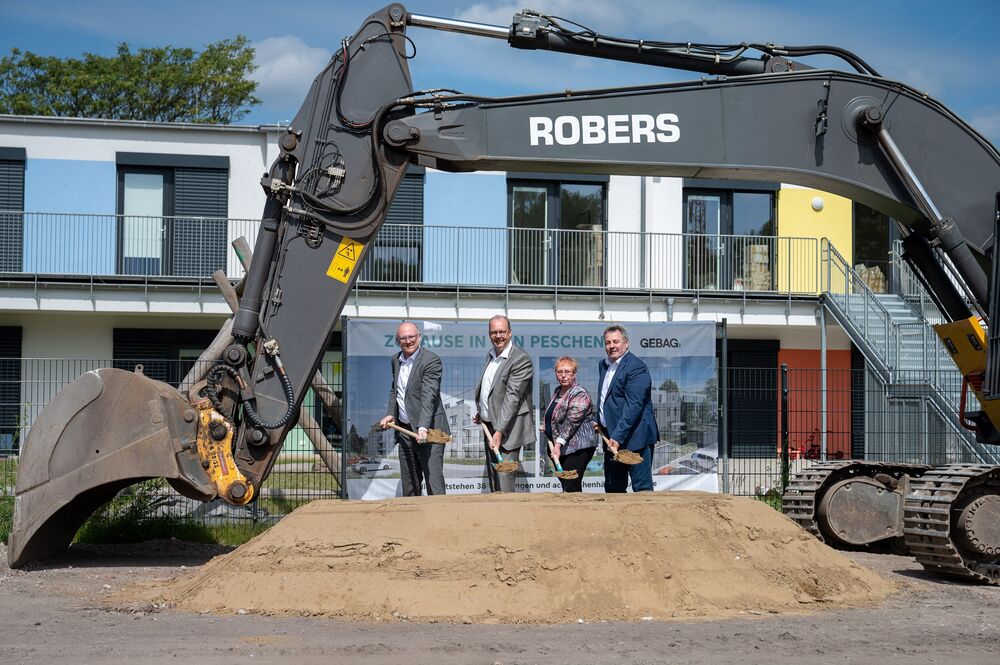 Eine Frau und drei Männer stehen in einer Reihe und führen mit einem Spaten den Spatenstich durch. Vor Ihnen befindet sich ein Haufen Sand. Neben Ihnen steht ein Bagger. Die Schaufel des Baggers ist über sie gebogen. Am Bauzaun hinter Ihnen hängt ein Banner, auf welchem das Neubauprojekt abgebildet ist.
