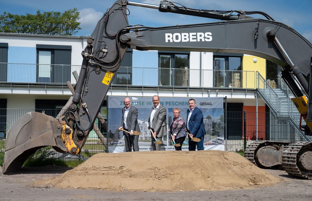 Eine Frau und drei Männer stehen in einer Reihe und führen mit einem Spaten den Spatenstich durch. Vor Ihnen befindet sich ein Haufen Sand. Neben Ihnen steht ein Bagger. Die Schaufel des Baggers ist über sie gebogen. Am Bauzaun hinter Ihnen hängt ein Banner, auf welchem das Neubauprojekt abgebildet ist.