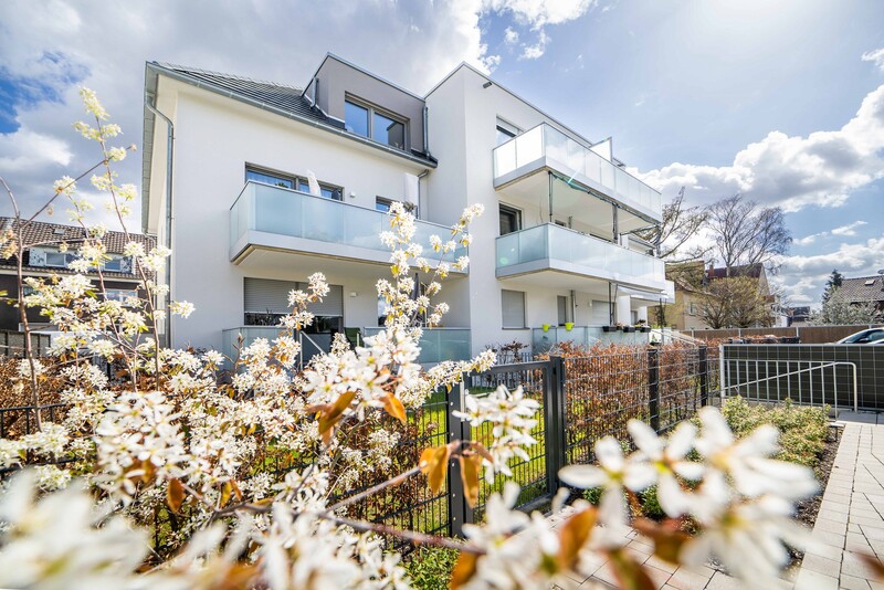 Blick auf ein weißes Mehrfamilienhaus mit Glasbalkonen vor blauem Himmel, Sonnenlicht von oben rechts. Im Vordergrund weiße Blüten.