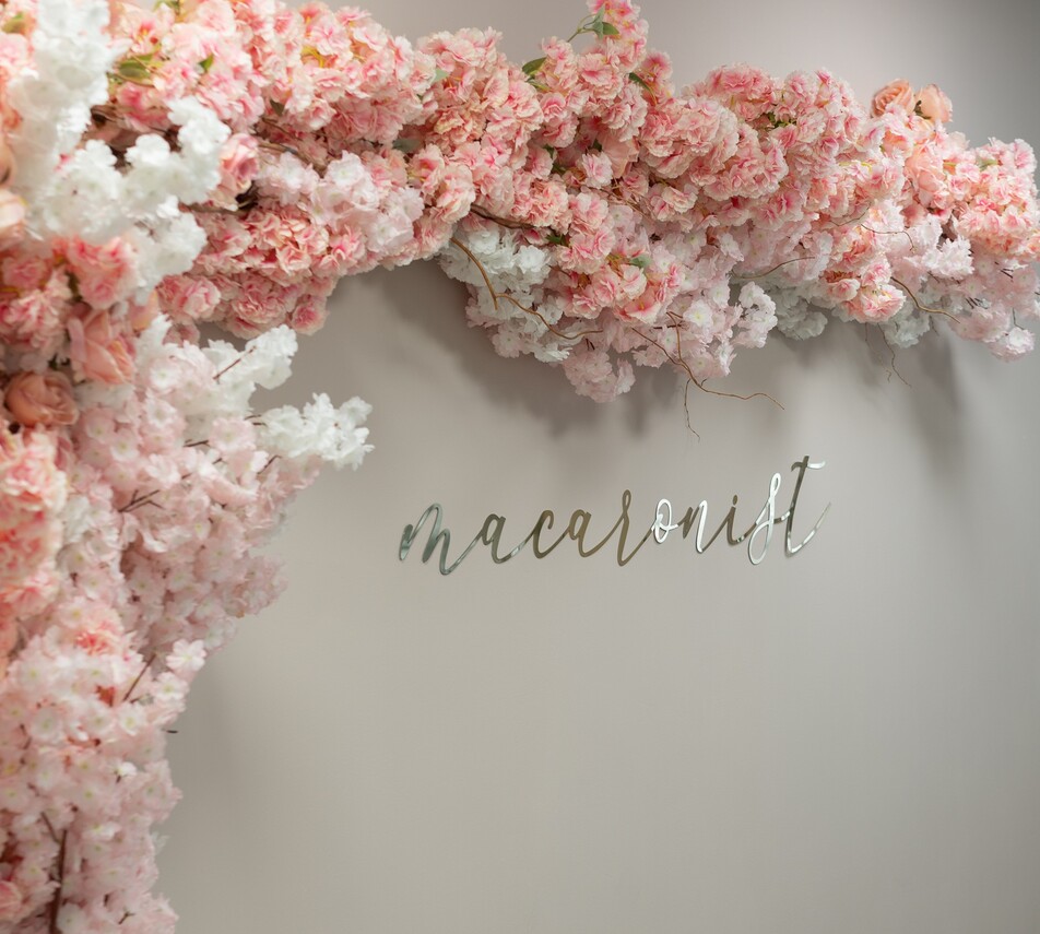 Rosa und weiße Blütenranken schmücken eine helle Wand mit dem Wort "Macaronist" in silbernen Buchstaben.
