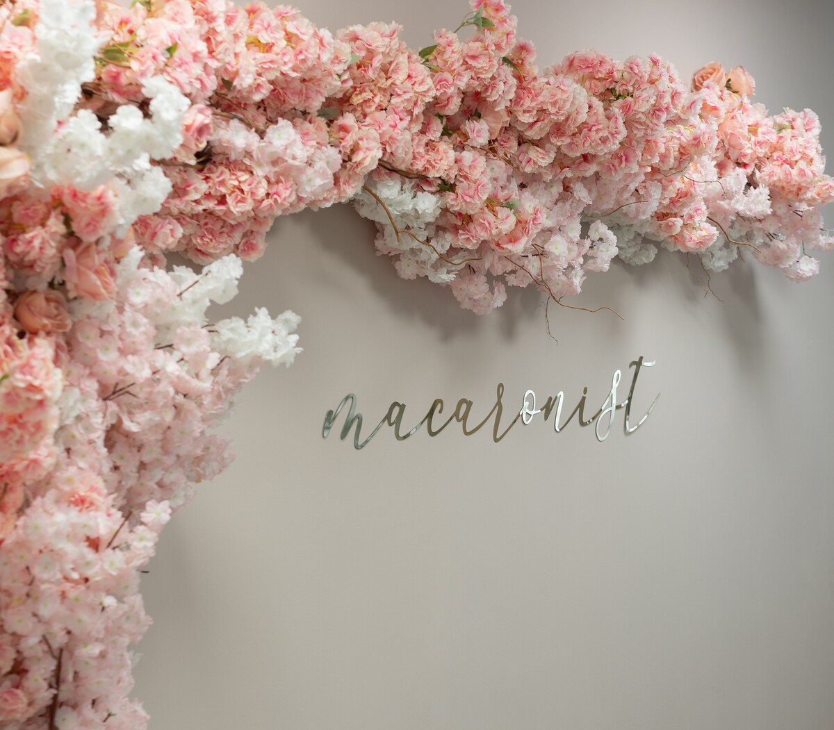 Rosa und weiße Blütenranken schmücken eine helle Wand mit dem Wort "Macaronist" in silbernen Buchstaben.