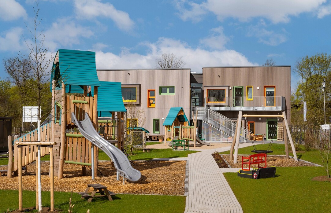 Moderner Spielplatz mit gepflastertem Weg und grünem Rasen. Im Hintergrund die Kita.