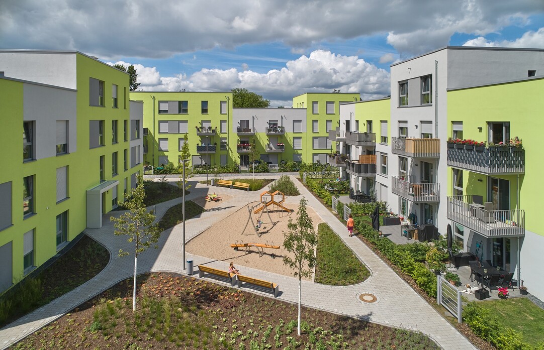 Blick in den Innenhof des Quartiers. Grün-weiße Gebäude gruppieren sich um einen Spielplatz, Wege, Bäume.