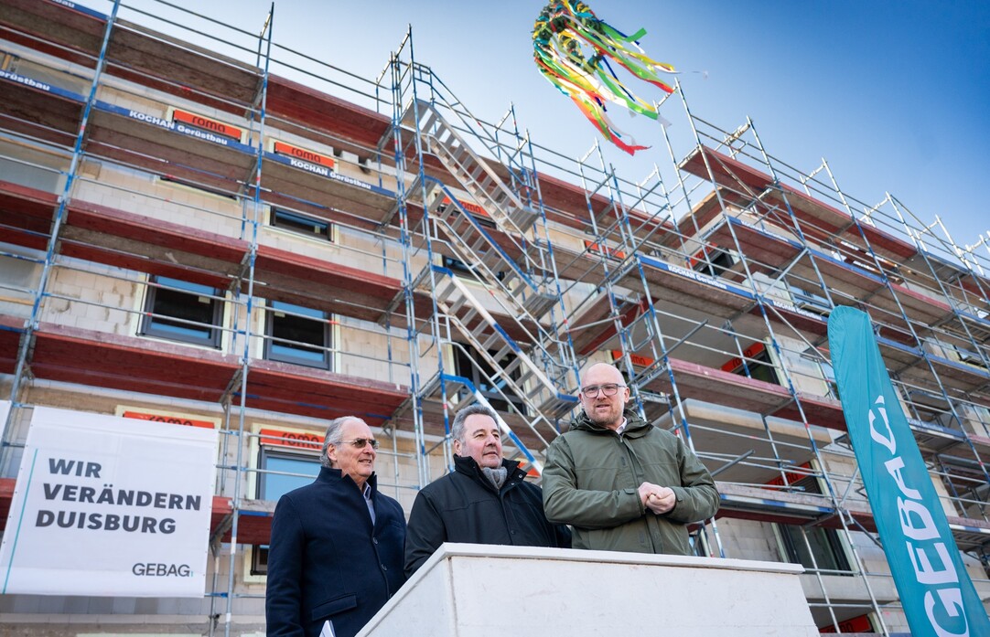 Drei Herren stehen auf einem Podest vor einem Neubau. Das Gebäude ist von einem Gerüst umgeben, an welchem ein Banner der GEBAG hängt. Neben dem Podest befindet sich ein Aufsteller mit dem GEBAG Logo. Über den drei Herren hängt der Richtkranz.