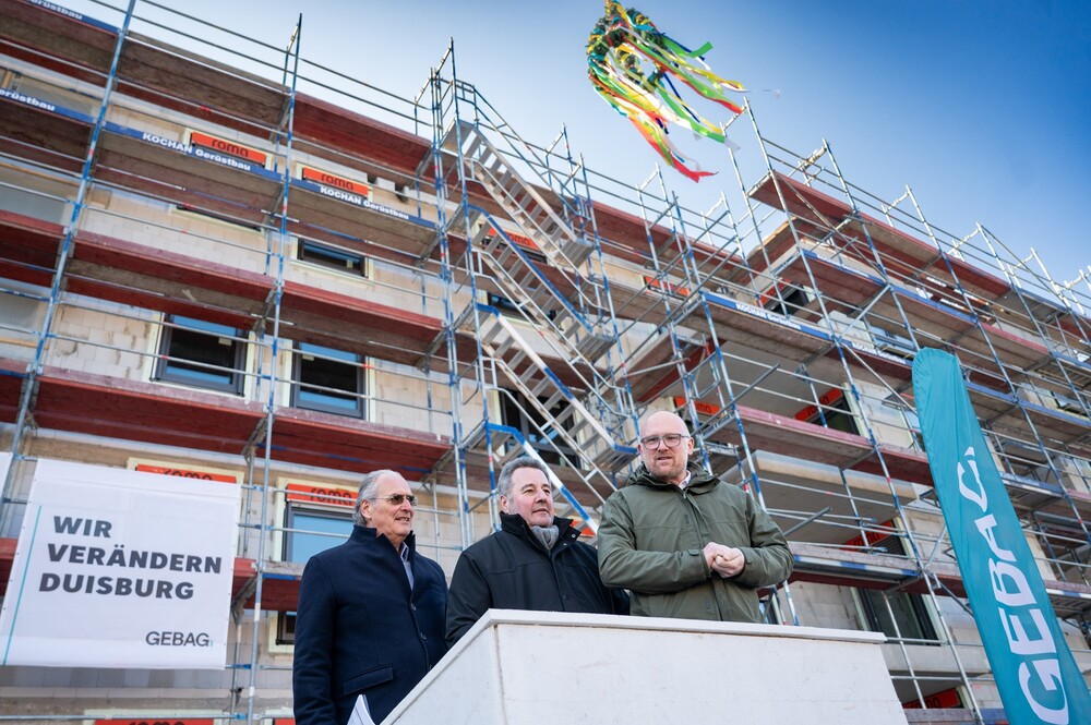 Drei Herren stehen auf einem Podest vor einem Neubau. Das Gebäude ist von einem Gerüst umgeben, an welchem ein Banner der GEBAG hängt. Neben dem Podest befindet sich ein Aufsteller mit dem GEBAG Logo. Über den drei Herren hängt der Richtkranz.