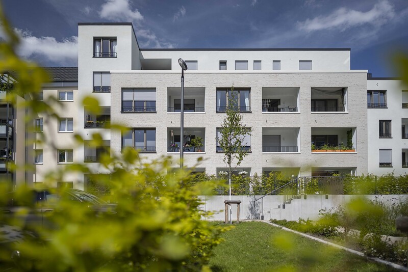 Im Vordergrund verschwommene Zweige, im Hintergrund der volle Blick auf den Neubau mit weißer Fassade und Loggien. 