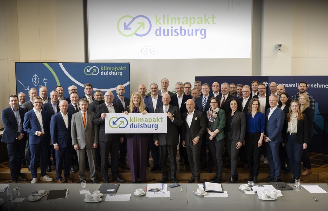Eine Gruppe mehrere Menschen steht zusammen und hält ein Schild mit dem Schriftzug "Klimapakt Duisburg" hoch.